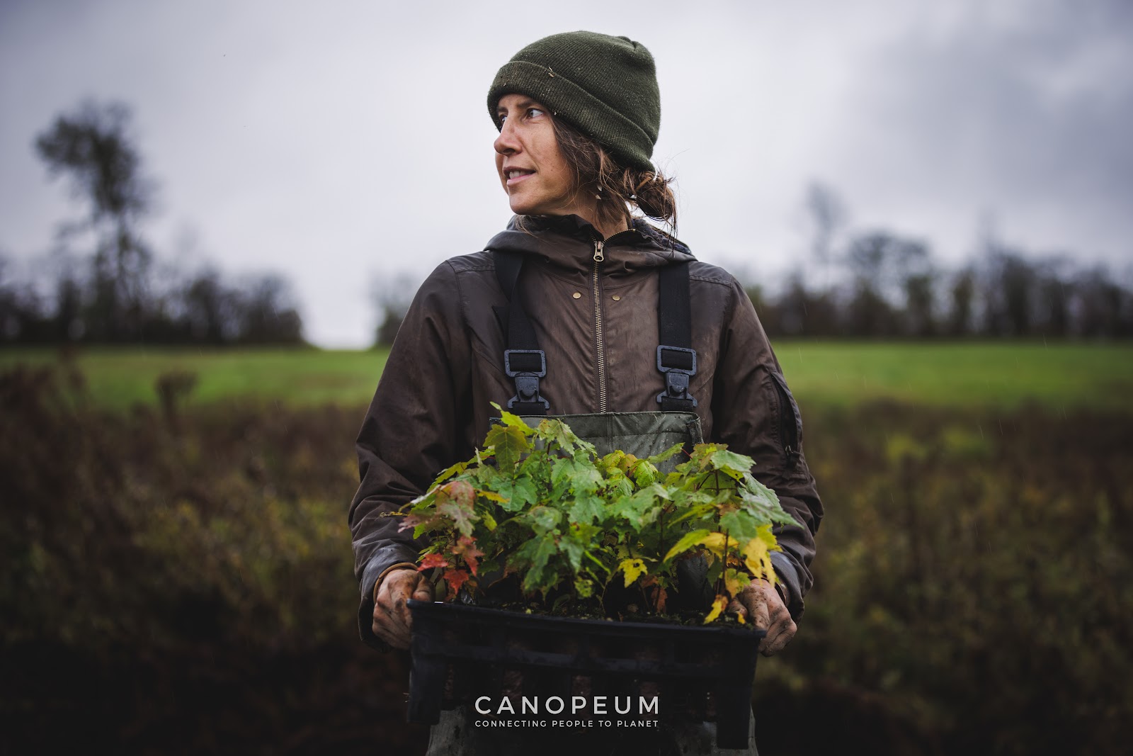 A person holding a tray of young trees outdoors, with the Canopeum name below.